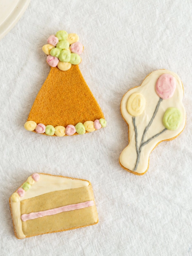 Overhead view of a cake slice, party hat and balloon bone Celebration iced dog biscuit trio on white fabric.