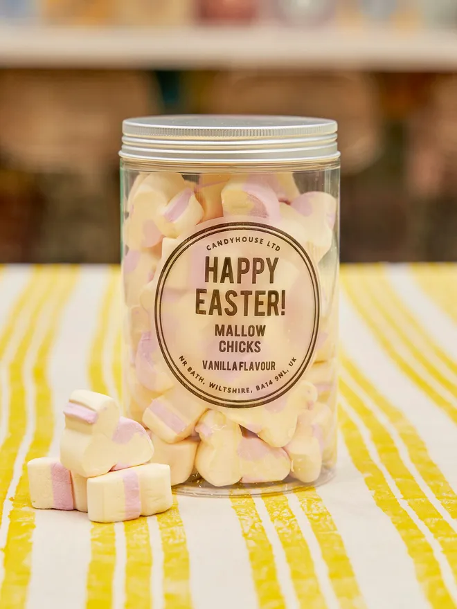 A clear jar full of mallow chicks placed on a yellow and white tablecloth with a few mallow chicks on the side 