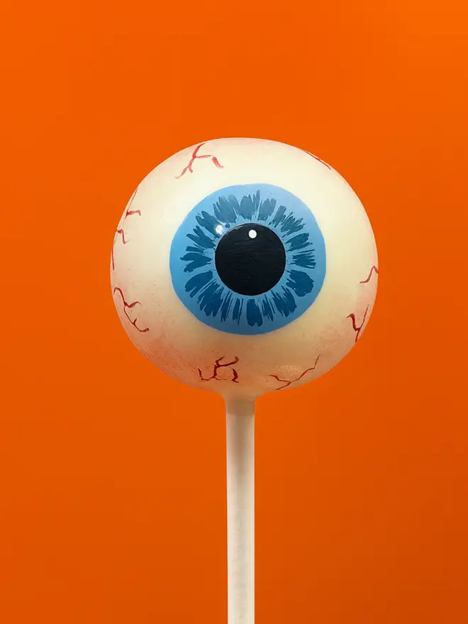 Realistic human eyeball cake pop on a white stick against a bright orange background
