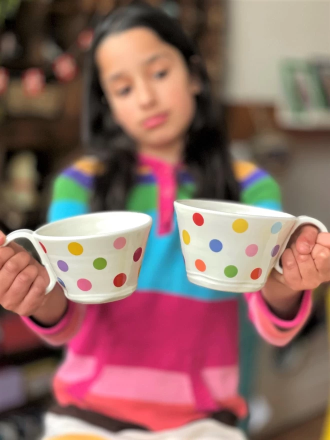 bemused chiild wearing a brightly coloured jumper , holding a large porcelain cup with polakdot pattern in each hand