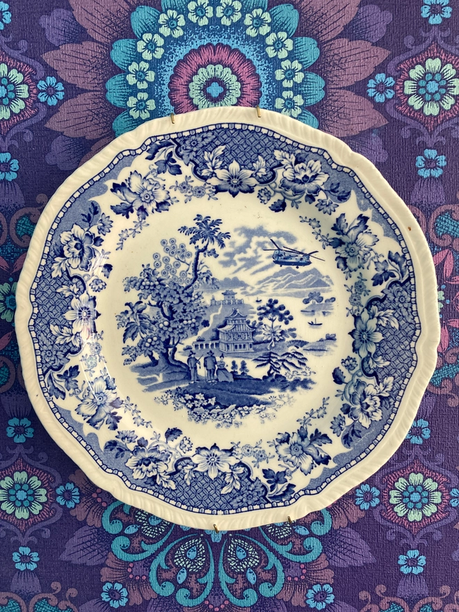 A Chinese style blue and white china plate portrays three people standing near a property. One is holding a fishing rod. A Chinook helicopter flies overhead.