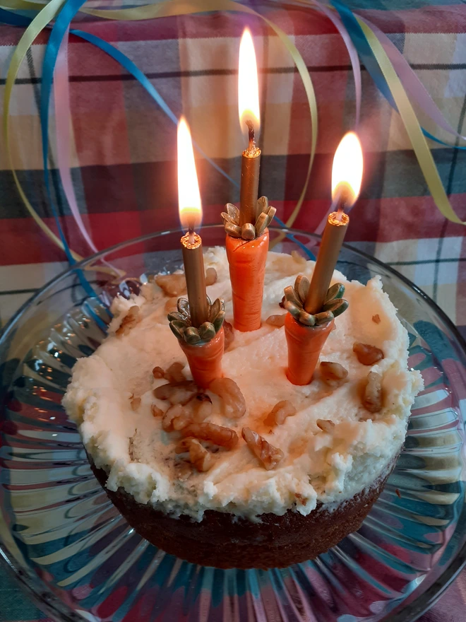 Three ceramic carrot candleholders placed in a frosted carrot cake holding lit standard birthday candles, styled with colourful ribbons in a festive spring setting.