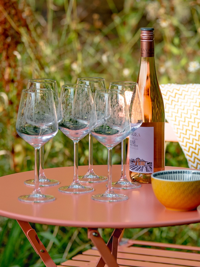 Meadow Wine Glass, a group of six etched wine glasses with wildflower patterns sit on a bright pink patio table next to a bottle of rosé wine and a small yellow bowl.