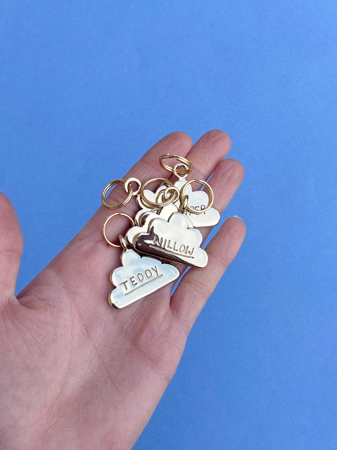 a hand holding a batch of shiny brass pet tags in the shape of a cloud. they each have pet's names engraved on the front with an underline.
