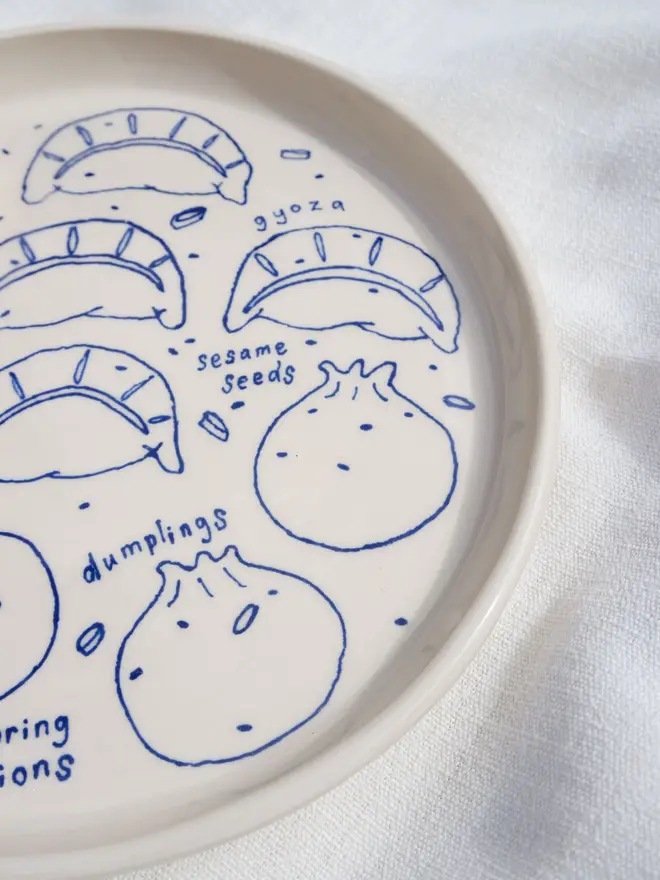 Dumplings Dippy Dinner Plate, a dipping plate sitting on a white bed cloth. It is covered in blue outlines for dumplings. 