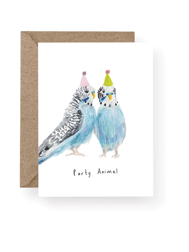 Budgie Party Animal Greeting Card, Card Has a White Base With Two Hand Painted Watercolour Budgies Wearing Party Hats.  Sitting On A Recycled Brown Kraft Envelope.  There Is Black Handwritten Text Underneath The Budgies Which Reads ‘ Party Animal’