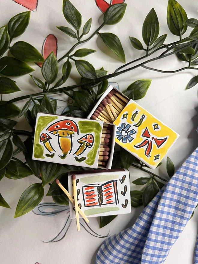 A whimsical arrangement featuring hand-painted matchboxes with vibrant designs, including mushrooms, abstract patterns, and a book.