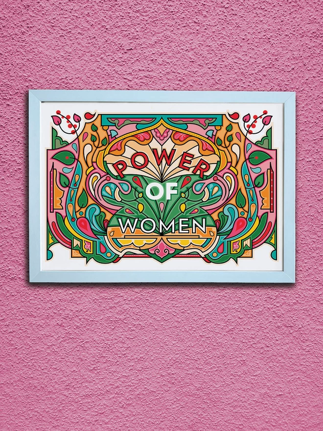 A vibrant, landscape illustration in greens, oranges and reds, with Power of Women written in the centre. It is in a white frame hung on a textured pink wall.