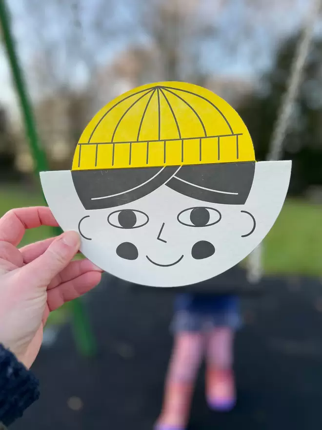 Hand holding a round shaped card with a childs face on and yellow hat. The card is being held in front of a childs swing.  