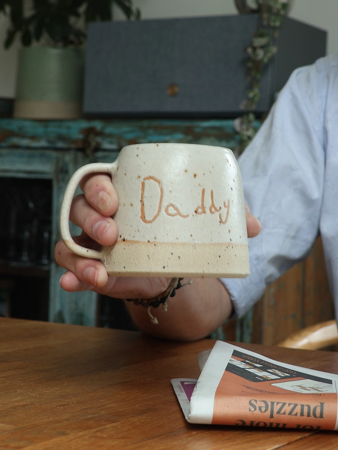 Personalised Kids' Handwriting Handmade Stoneware Mug held above table with the name 'Daddy'