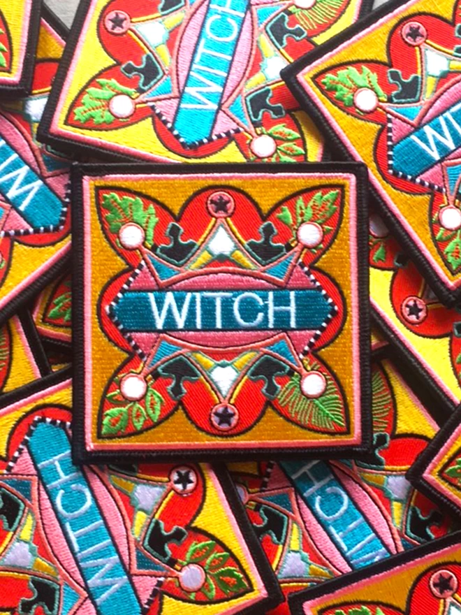 The patch sits on a scattered pile of many other patches. At the centre of the patch is the word “Witch” embroidered in white on a light blue strip. Surrounding this is a colourful, bold design which sits on a yellow background, with a thin pink inner border and a thick black outer border.