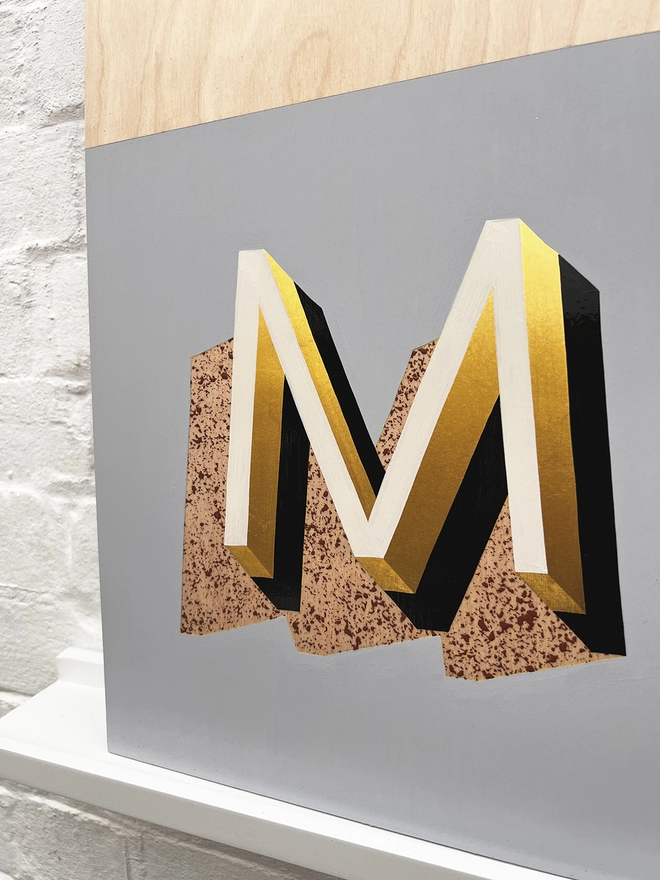 Close up of letter M from HOME hand painted sign in 23 carat gold leaf, pale blue, off white, tan, against a white brick wall, at an angle. 
