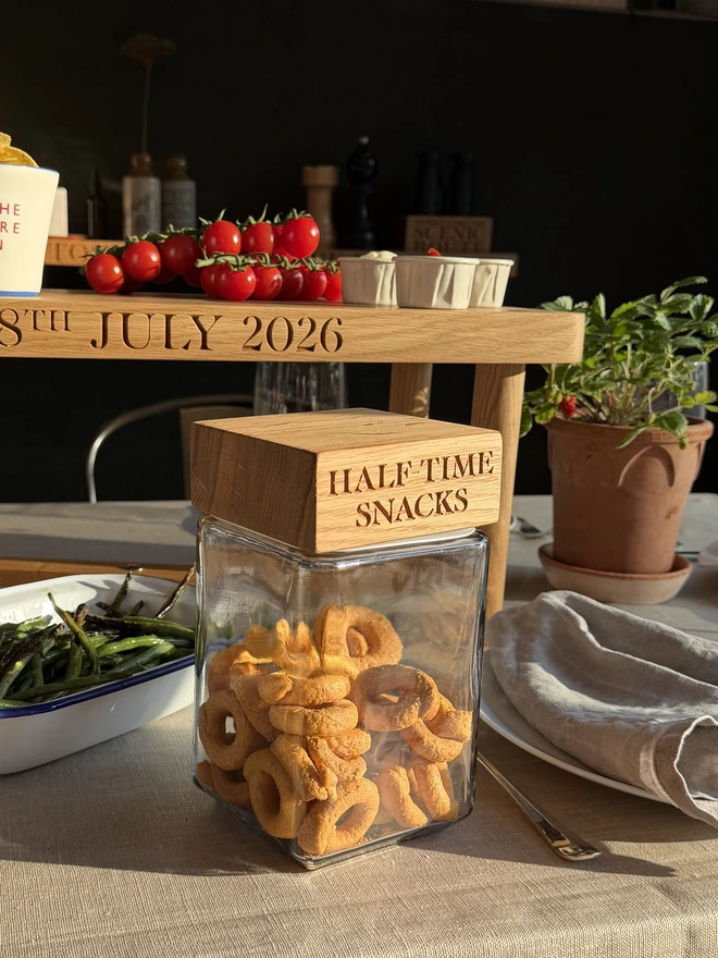 Personalised 'Half-Time Snacks' oak jar with engraved oak lid, filled with snacks and styled on a dressed table with fresh tomatoes and a potted strawberry plant.