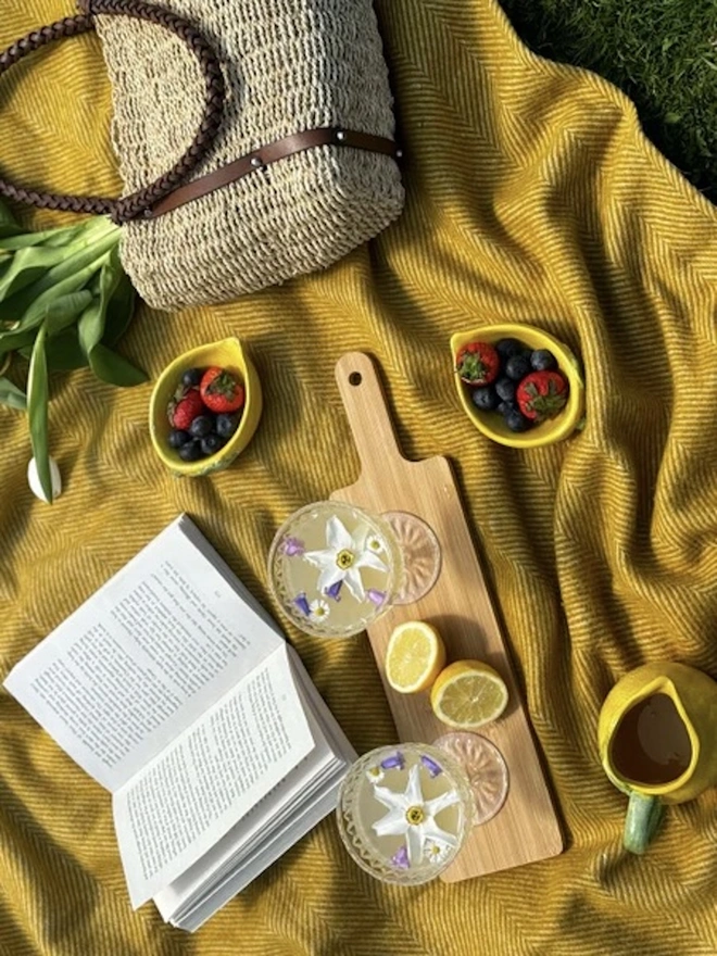 Mustard Recycled Wool Tartan Blanket, a yellow picnic blanket sitting outside on the grass. It is accompanied by a wooden hamper full of food and a book. 