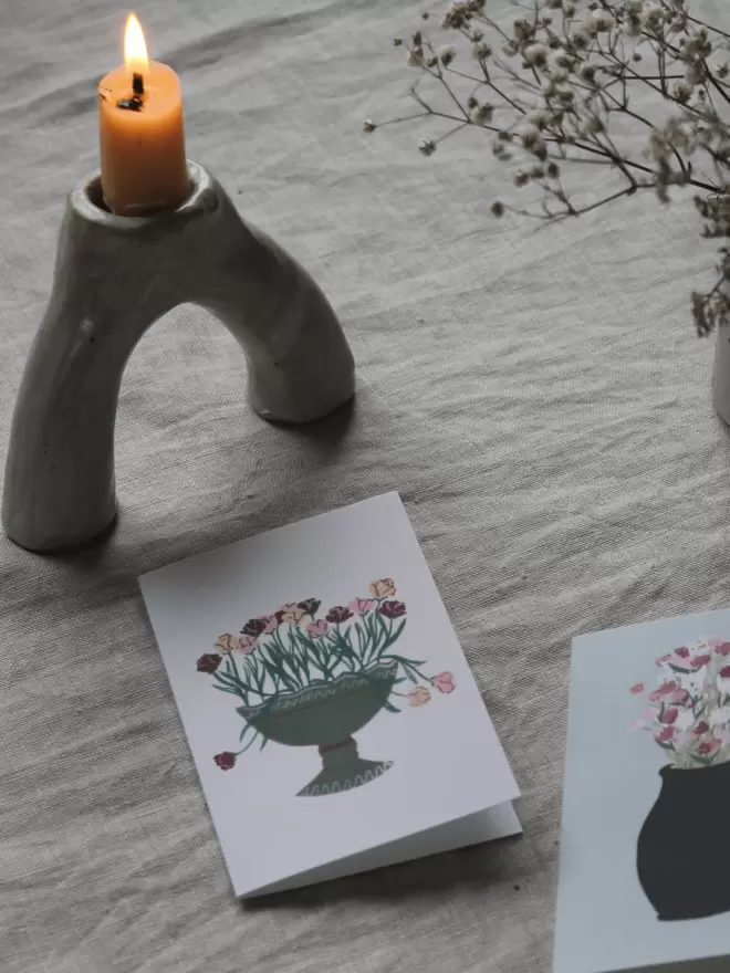 card with green vase with flowers arranged in it, on linen tablecloth next to a lit candle in a holder 