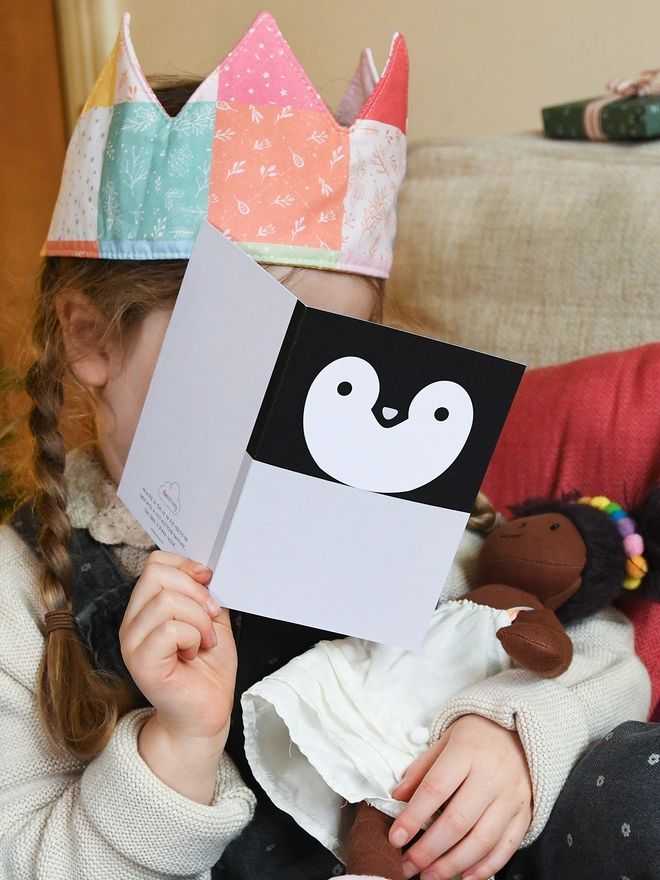 A child wearing a patchwork crown holds a penguin greetings card in front of them.