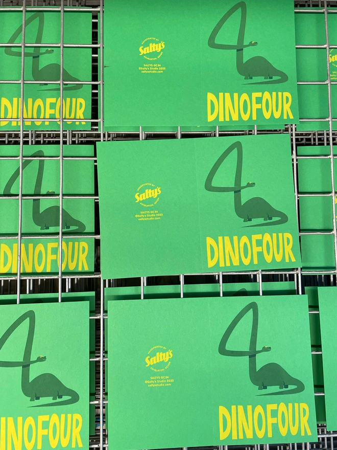 Several bright green cards on a screen printing drying rack. Each card has the number four that has been created by the very long-necked, happy dinosaur. The words DINOFOUR are at the bottom in bright Yello type