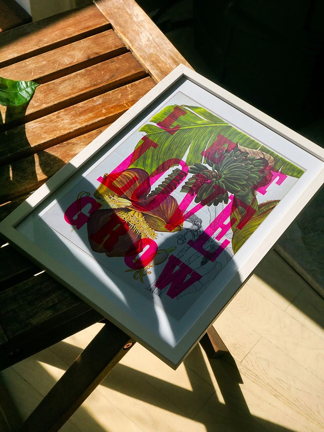 A framed limited edition artwork rests on a wooden surface, partially lit by sunlight. The letterpress print features a mix of bright pink and green leaves with the phrase "LET LOVE GROW" in bold, colorful letters rendered in fluorescent ink.