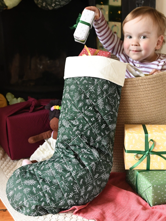 A young toddler wearing striped pyjamas sits in a wicker basket and pulls a present out of a green quilted Christmas stocking.