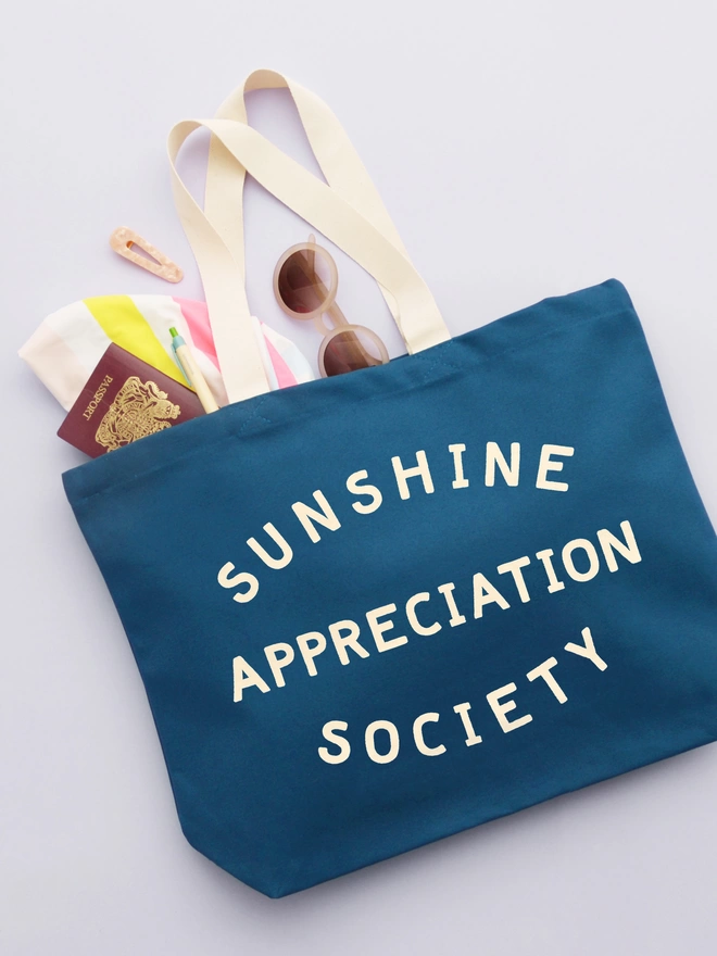 a large blue canvas tote bag with the words sunshine appreciation society on laying on a purple background with sunglasses, a passport and swimsuit spilling out