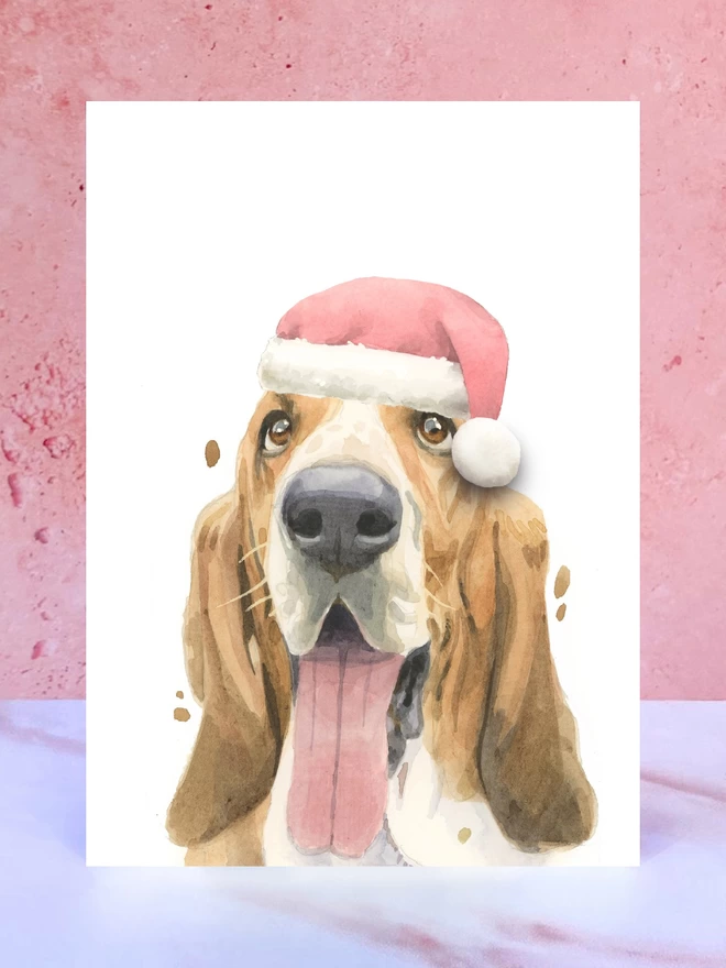 A Christmas card featuring a hand painted design of a basset hound, stood upright on a marble surface surrounded by pompoms. 