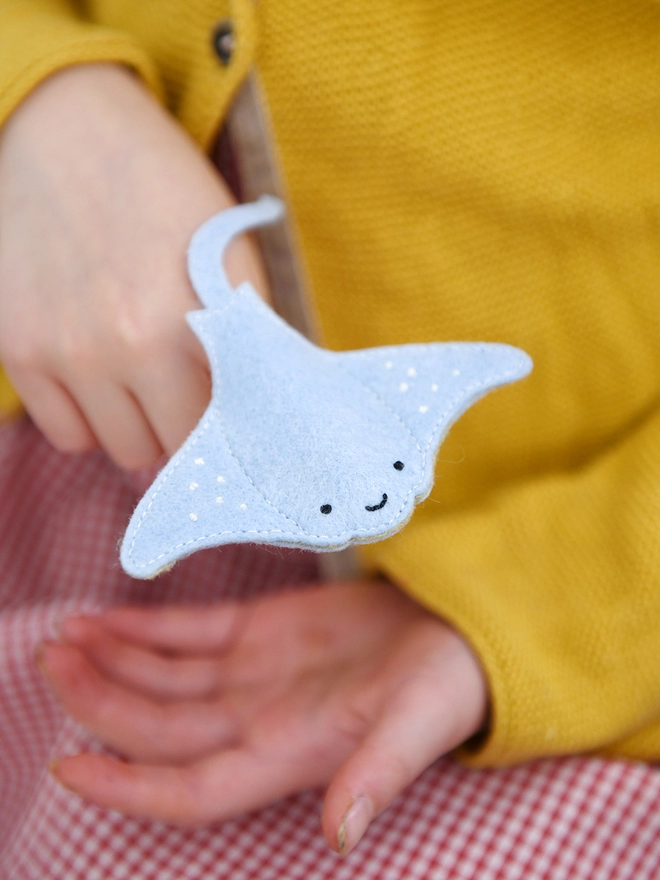 A young child wearing a yellow cardigan is holding a soft blue felt manta ray finger puppet on their fingers.