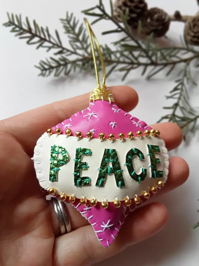 A Hetty and Dave pink hand stitched leatherette retro inspired bauble with green metallic lettering spelling out PEACE on a white band, adorned with tiny gold beads