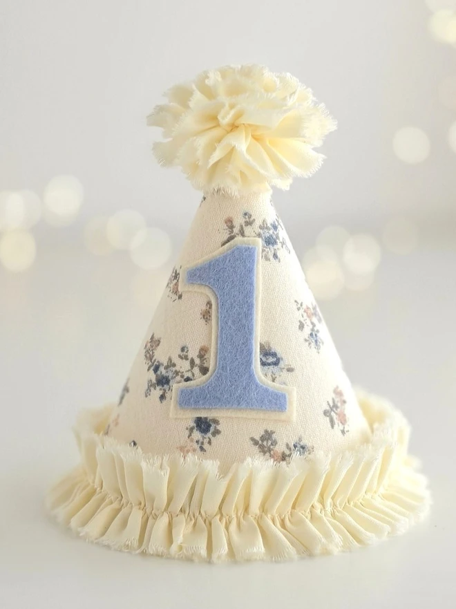 Handmade first birthday party hat in vanilla floral cotton with blue number one applique and soft gathered cotton trim.