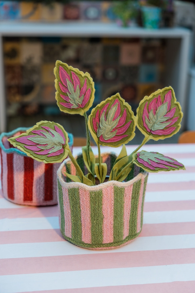 Pink and green striped scalloped jute basket on a pink and white striped tablecloth with a felt house plant sat inside