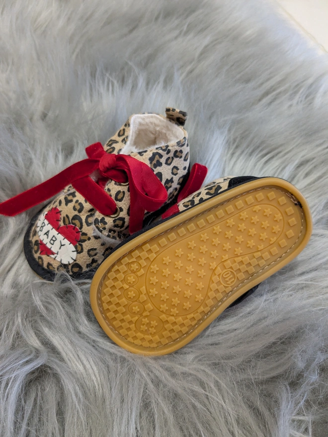 A pair of leopard print baby booties, lined with fur and with red velvet ribbon laces. A red heart with a white scroll adorns each booty. One says BABY, the other, LOVE in black stitches