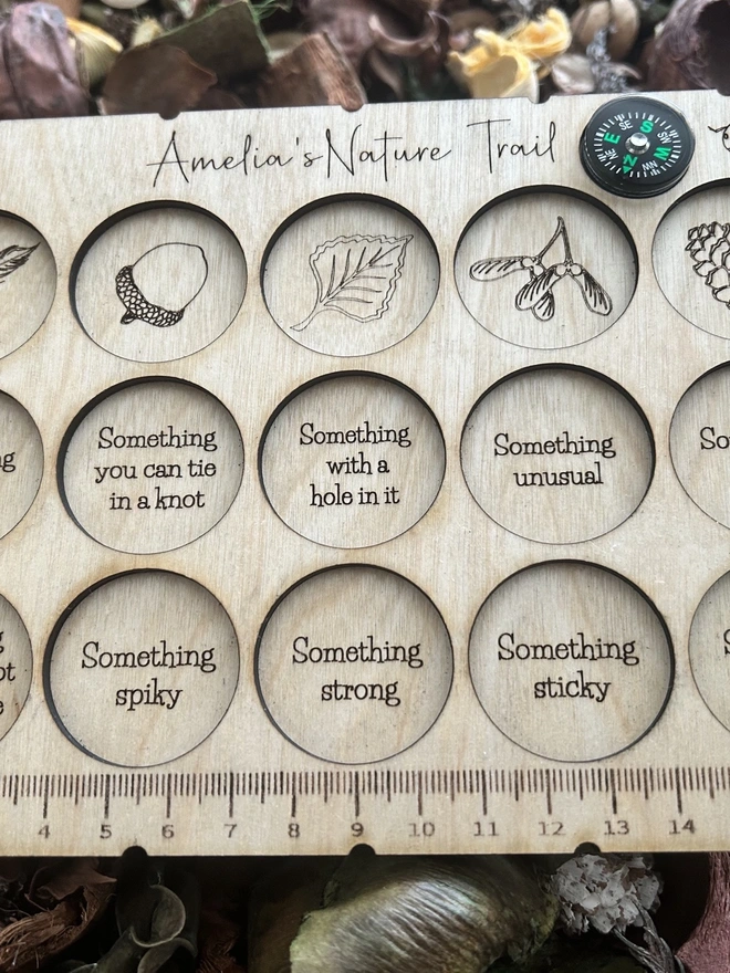 A personalised wooden nature scavenger hunt board displayed on an outdoor surface. The board is rectangular with rounded corners and engraved with 15 small collection slots held by clear bands. A custom name is engraved at the top, and a button compass with a wrist lanyard is attached for carrying. The wood has a smooth, light finish, and no items are placed in the slots; only the board is shown.