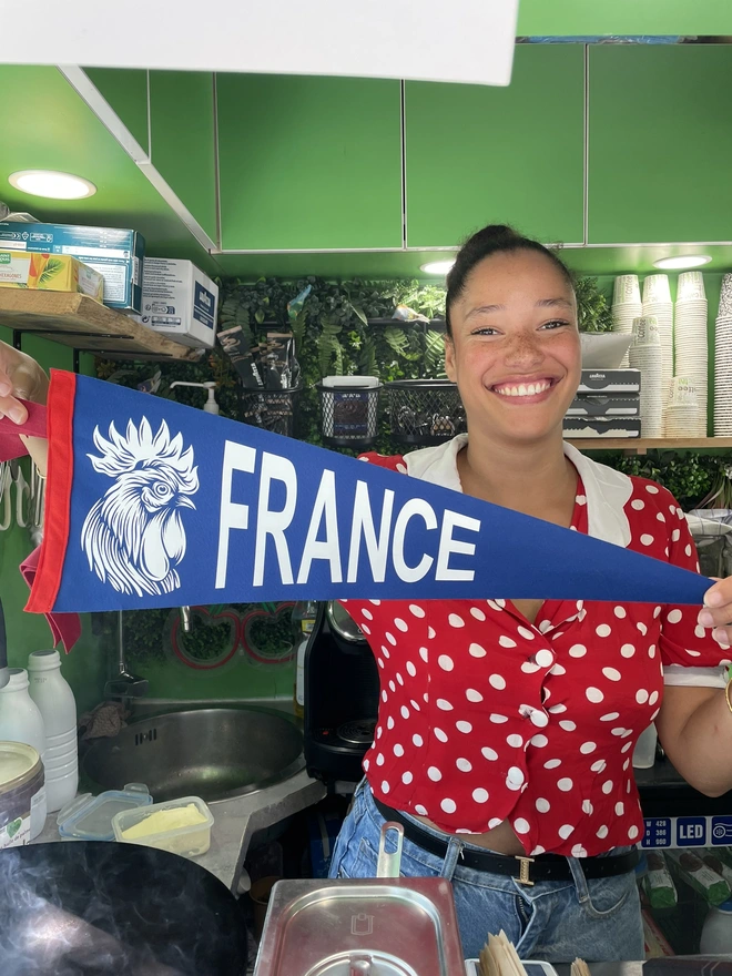 a smiling French girl in a cafe holds up a red white and blue felt pennant flag with the image of a rooster head (france's national bird) and the word France in bold