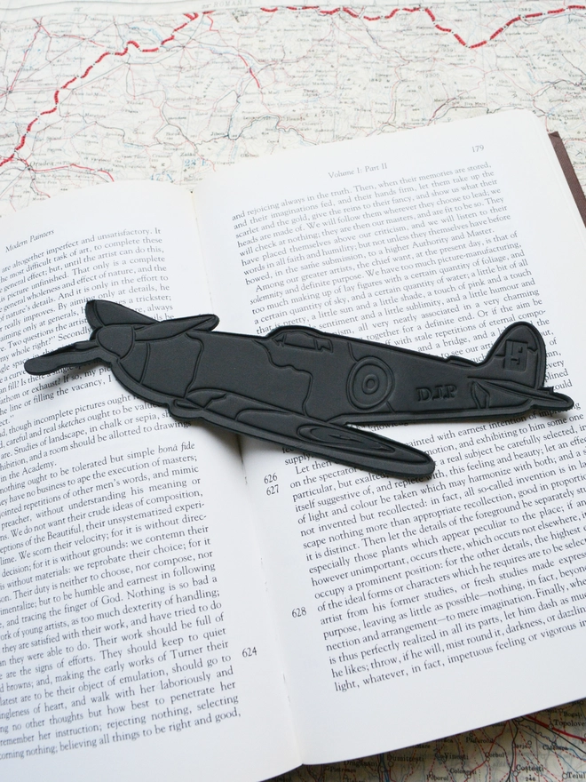 a black blind embossed leather bookmark of a ww2 spitfire airplane from the no.19 squadron with personalised initials in a reading book with a map by natthakur
