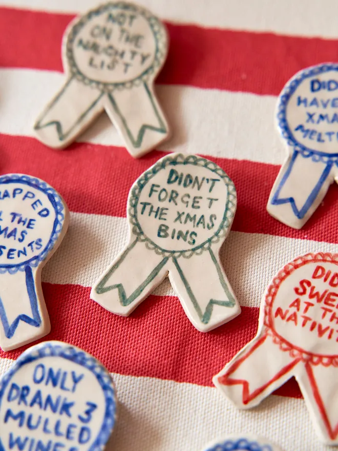A selection of Red, Green & Blue Handmade Ceramic Christmas Award Badges, handpainted with different phrases, atop a striped red and white tablecloth