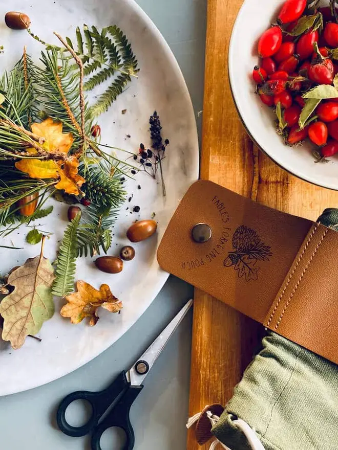 Personalised Foraging Drawstring Canvas Pouch, a green canvas pouch with a leather flap that reads "FABLE'S FORAGING POUCH" sits next to bowls of leaves, acorns, and red berries.