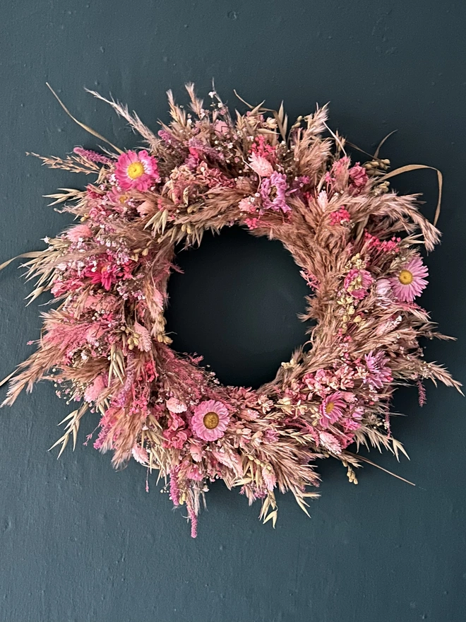 Pink Dried Floral Wreath. a circular wreath made of pink dried flowers hung up on a forest green wall.