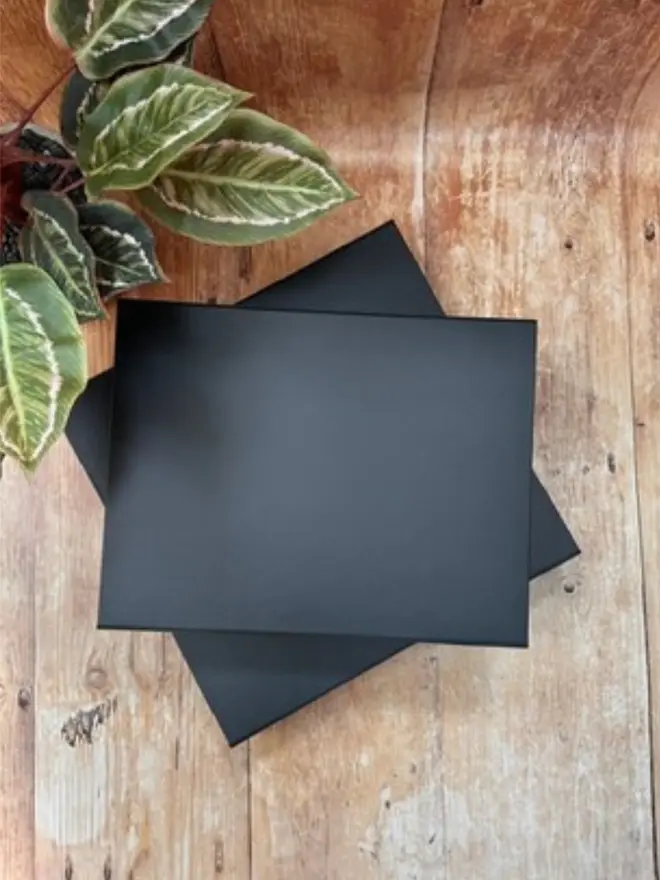 A pile of navy presentation boxes for photo albums or guest books is resting on a wooden work surface, next to a pot plant.
