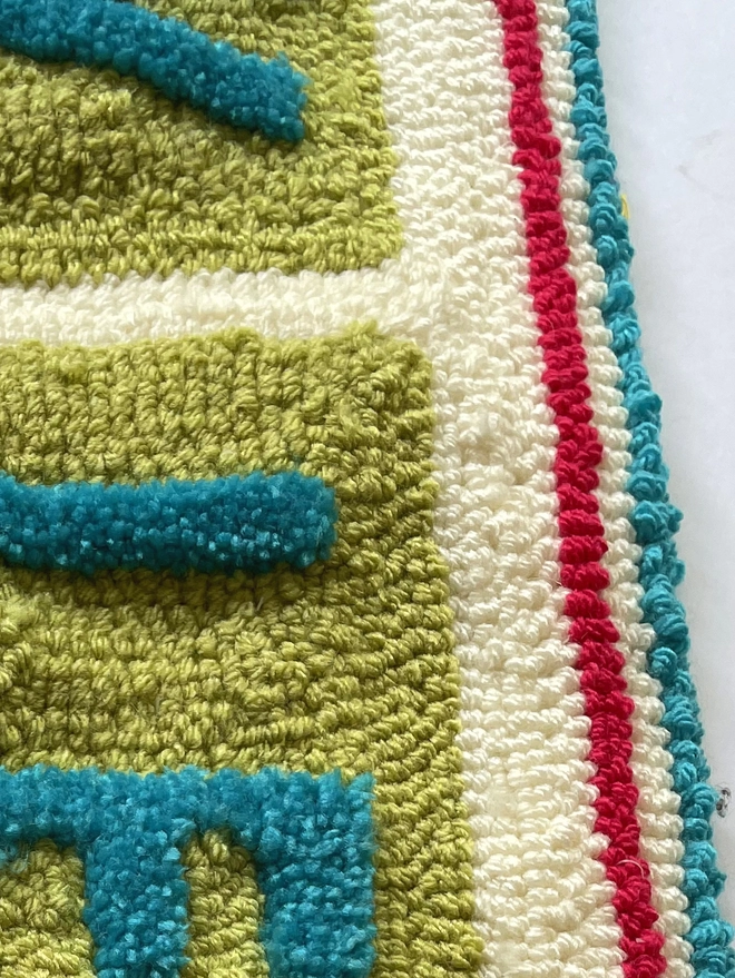 close up of blue and green wool loops forming the letters and red and blue stripes