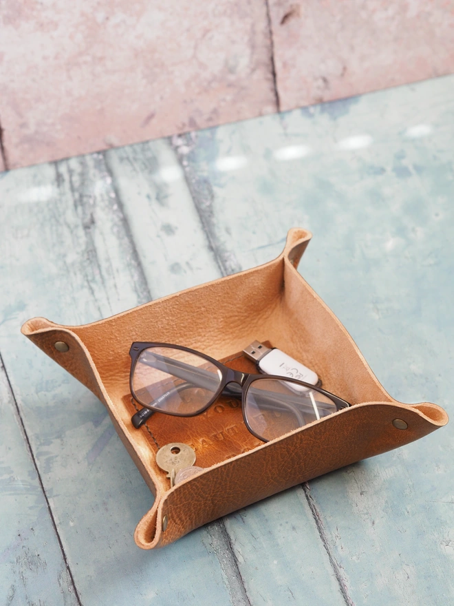 Personalised message leather coin tray. Hand dyed tan brown leather tray with a square stitched to the base which is personalised with a short but sweet message. Inside the tray is a pair of reading glasses, keys and a USB stick.