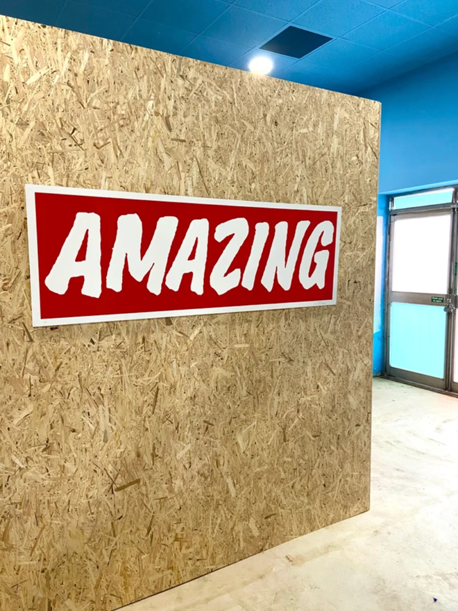 Very Big Amazing. The print seen against a wall. The design is a simple red background with a white, all caps AMAZING written.