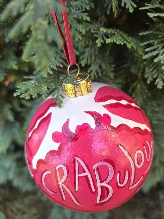 a bauble hanging in a christmas tree, hand painted with a crab design in dark pink and neon pink with the slogan 'Crabulous'