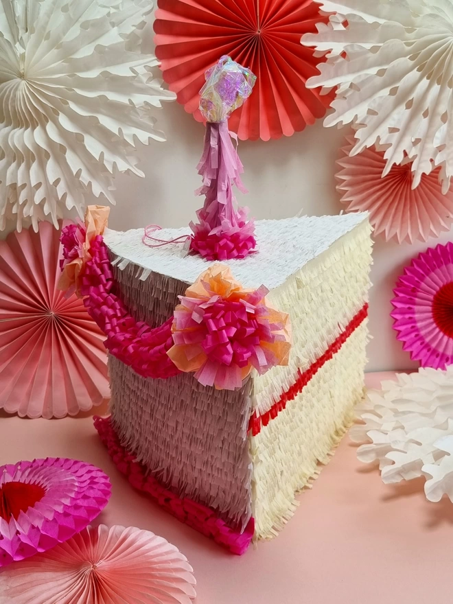 birthday cake slice pinata with white and pink icing and pink candle on a background of pink paper fans