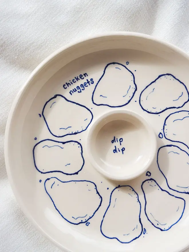 Snack Chicken Nuggets & Dip Plate, a white snack dish sitting on a white bed cloth. It is covered in blue outlines for chicken nuggets. 