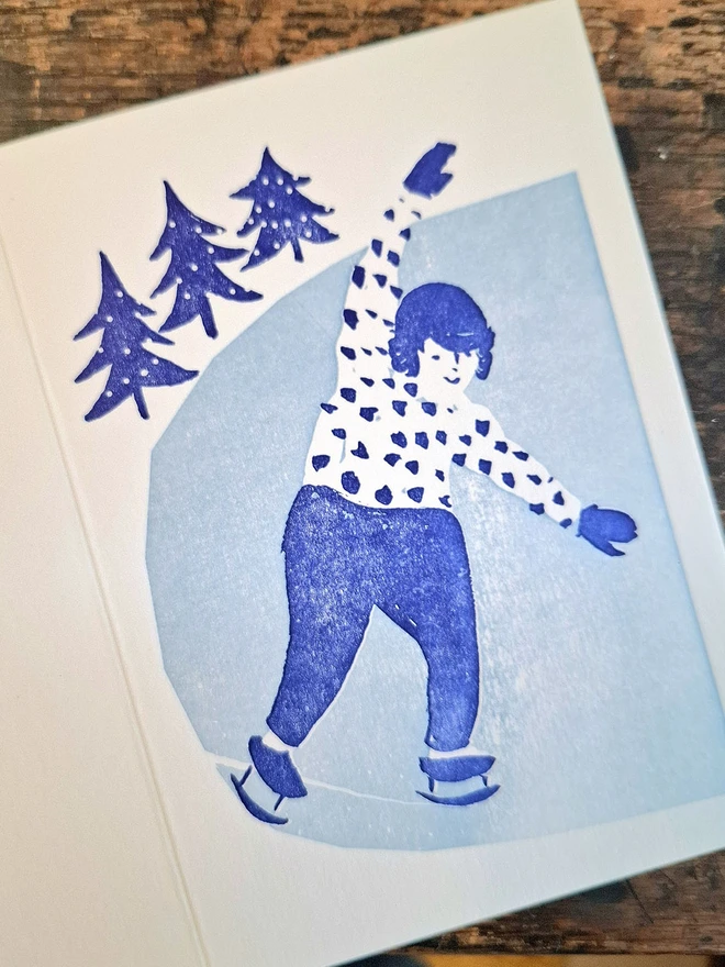 A greetings card lying open with the image face up.  The illustration on the front of the card is a handprinted image of a smiling girl iceskating, printed in light blue and dark blue.