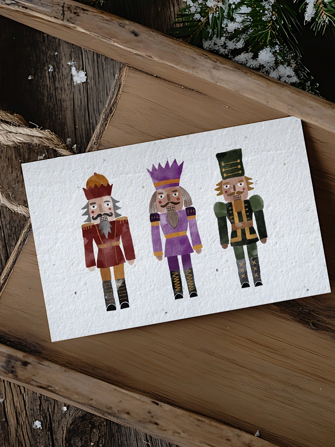 A Christmas card featuring three watercolour nutcracker. The card is placed in a wooden sleigh in a Christmas scene.

