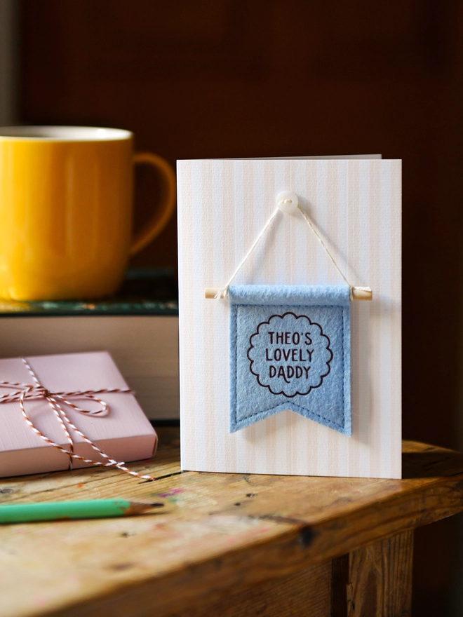Handmade Father’s Day card with personalised blue felt banner “Theo's Lovely Daddy”, miniature hanging flag keepsake on wooden table. The flag is hung on the card from a tiny mother of pearl button. Beside the card is a book, a mug, a wrapped gift.