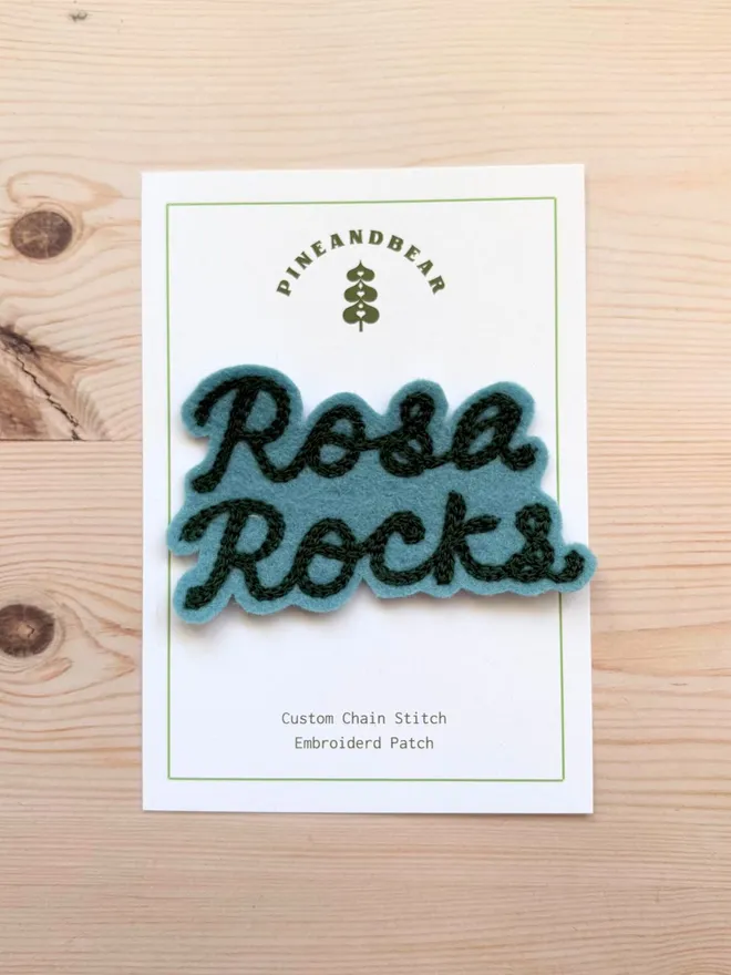 A chain stitched embroidered patch in teal and green spelling the word Rosa Rocks, on a white presentation card