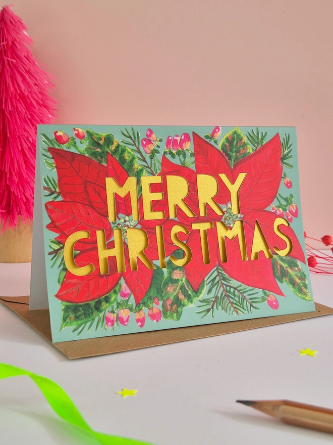 Red Poinsettia Illustration Christmas Card with green background, paper cut text that says Merry Christmas and has a pearlescent gold backing paper 