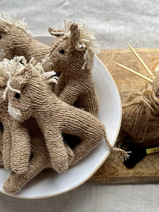 Hand Knitted Natural Dyed Wool And Alpaca Yarn Donkey Gift, a group of hand knitted donkeys sitting in a white bowl on a wooden chopping board on a table cloth. They are accompanied by some hey and some knitting accessories. 