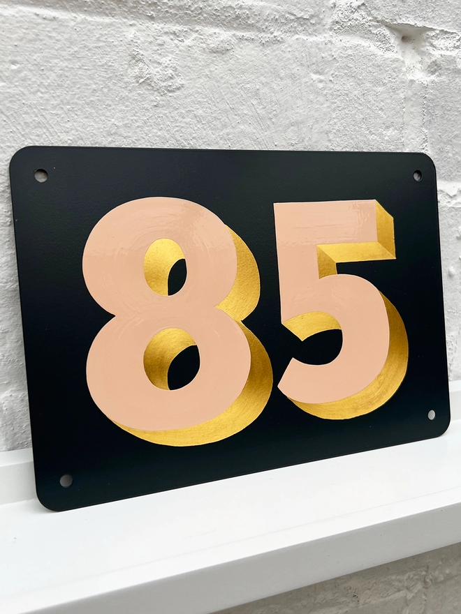 Hand painted peach and gold leaf house number 85 on an anthracite grey metal plaque against a white brick wall. 